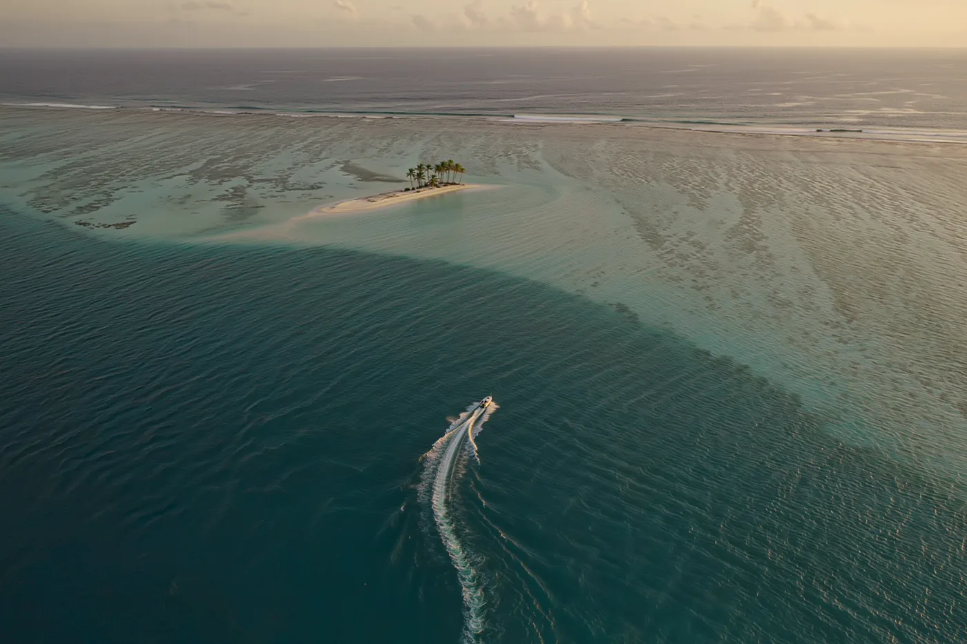 A speedboat crossing a turquoise lagoon towards a small palm island at golden hour