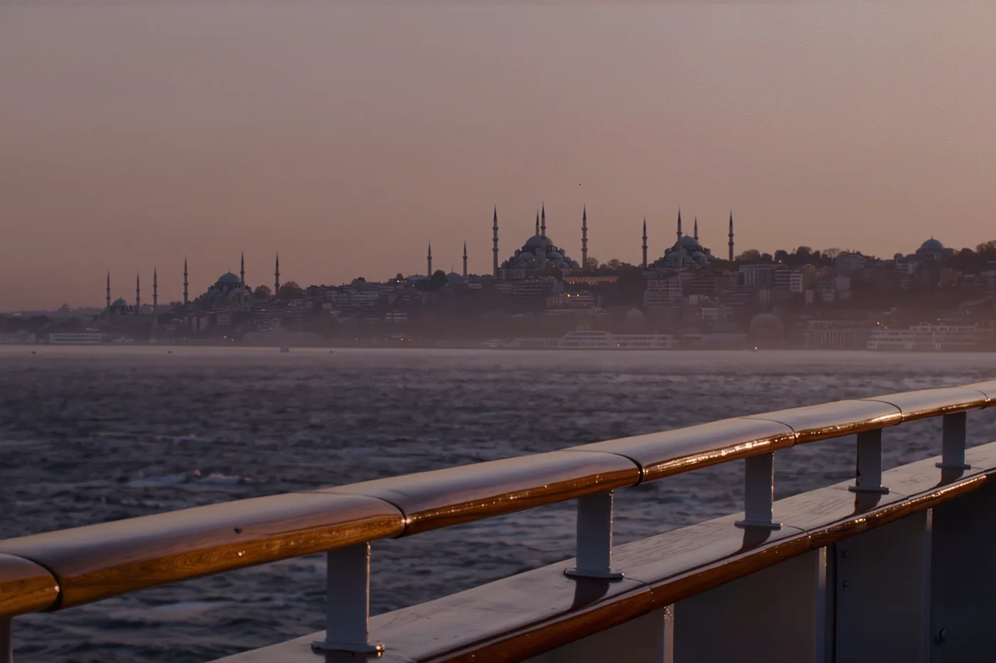 The Istanbul skyline at dusk seen across the water from a ship's deck railing